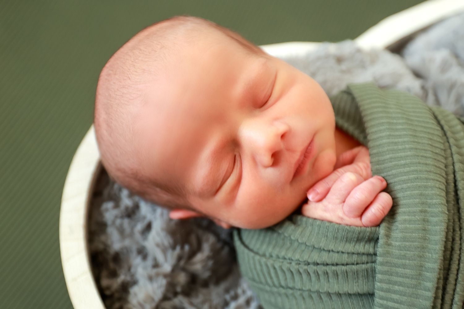 Peaceful newborn baby sleeping swaddled in green wrap inside a white bowl with gray fur lining.