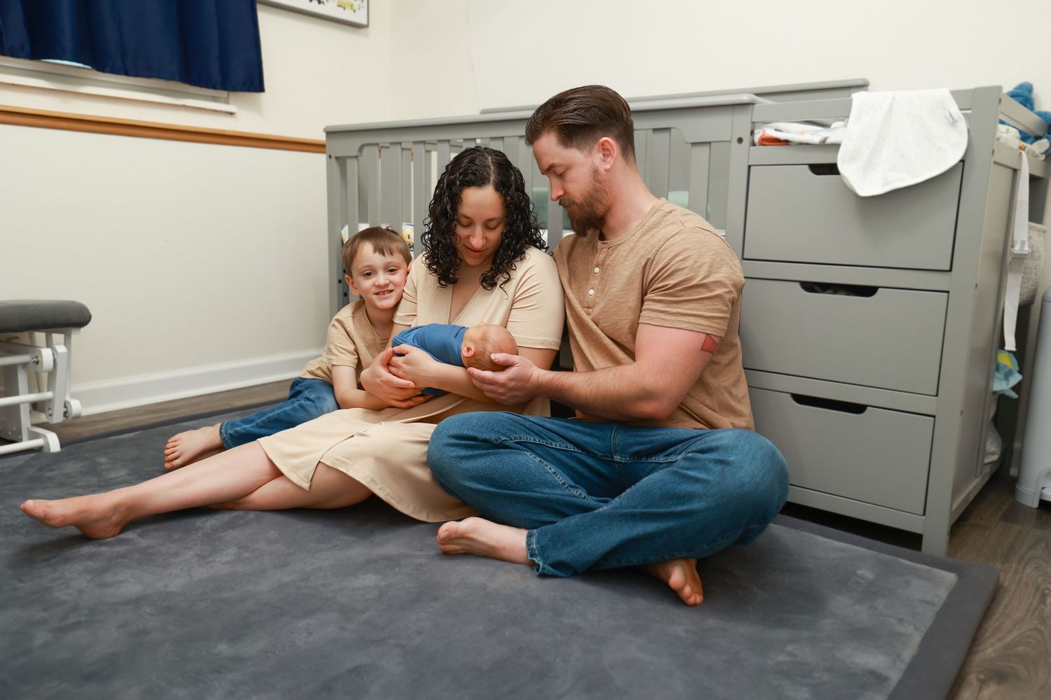 Happy family with young daughter sits on nursery floor admiring their swaddled newborn baby together.