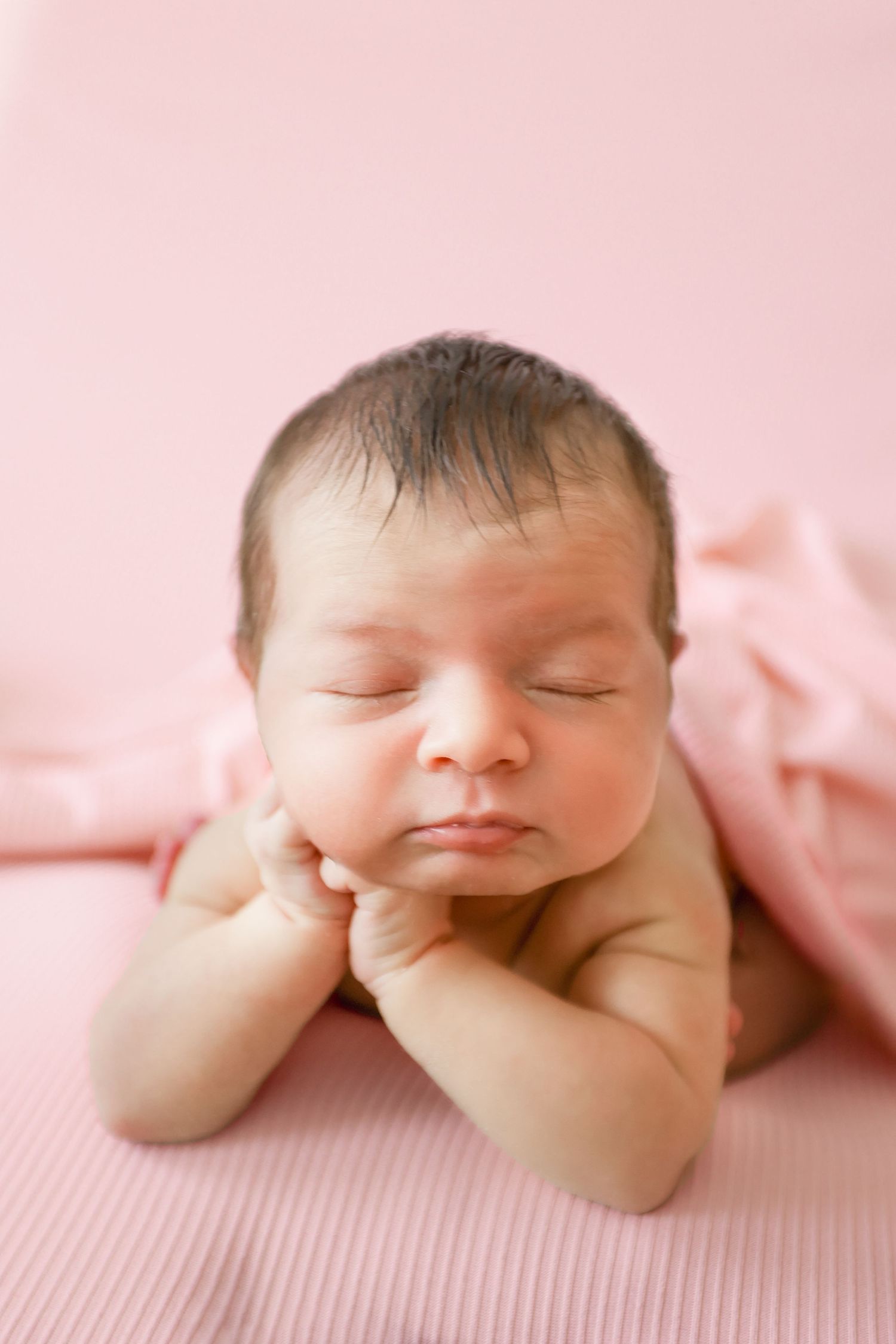 Close-up of sleeping newborn baby resting chin on hands, draped in soft pink blanket on pink background.