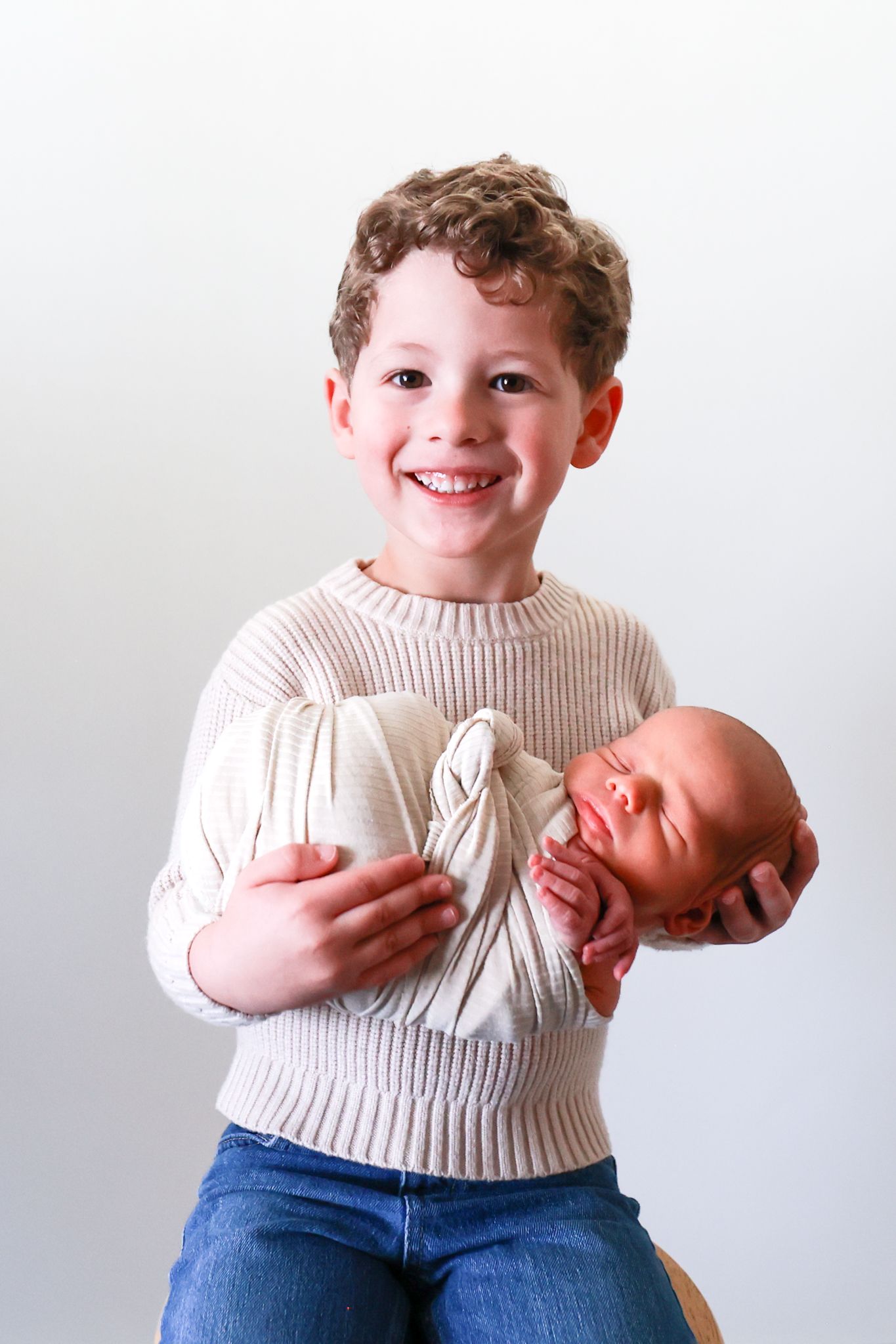 Smiling boy in beige sweater proudly holds sleeping newborn baby wrapped in white cloth against light background.
