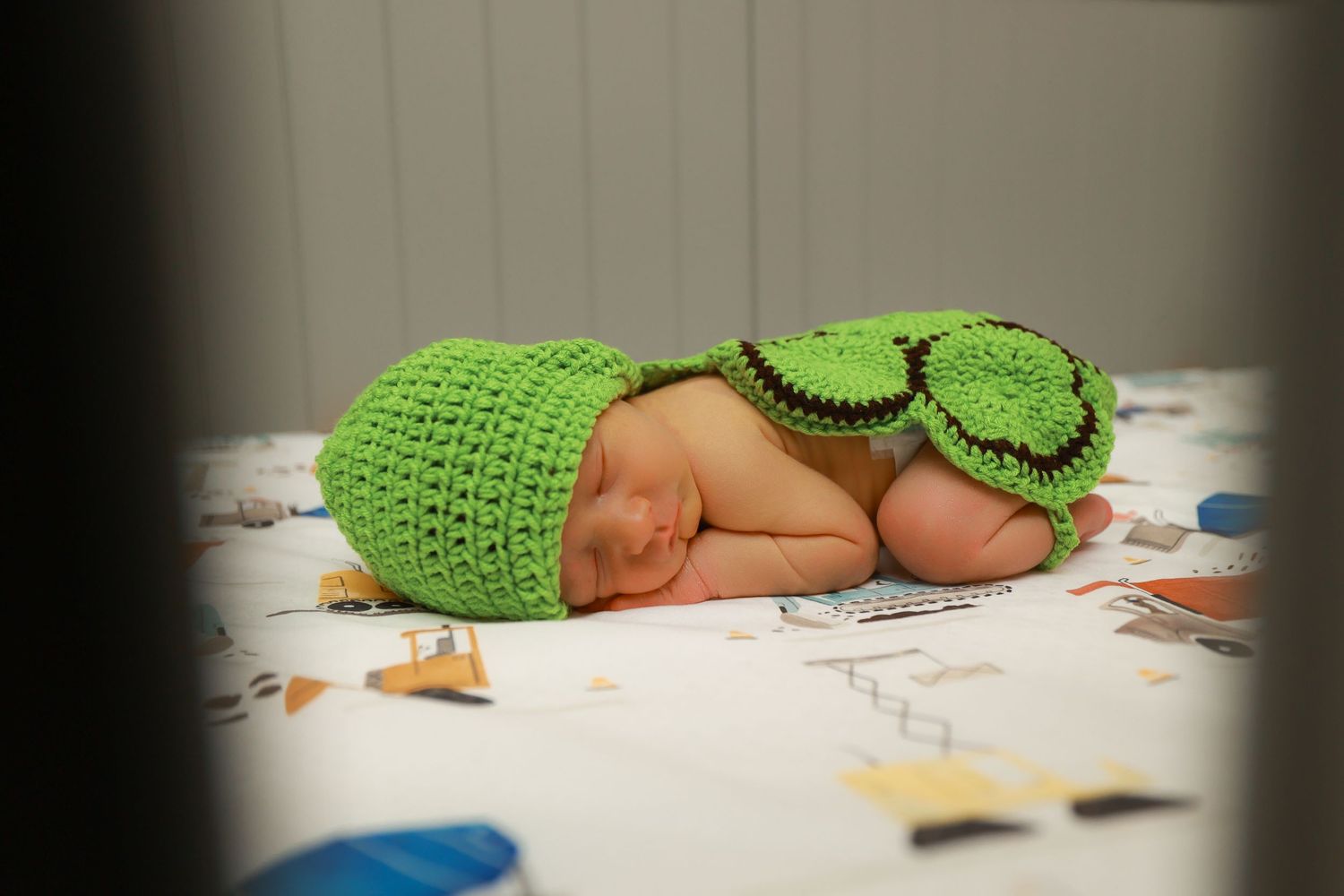 Newborn baby in green crocheted turtle outfit sleeping on a patterned blanket.