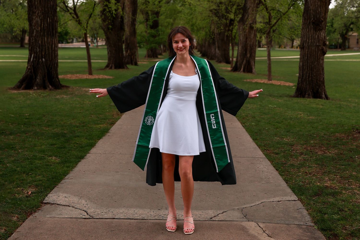 Young woman in white dress wearing green graduation stole and black gown, posing outdoors on a tree-lined path.