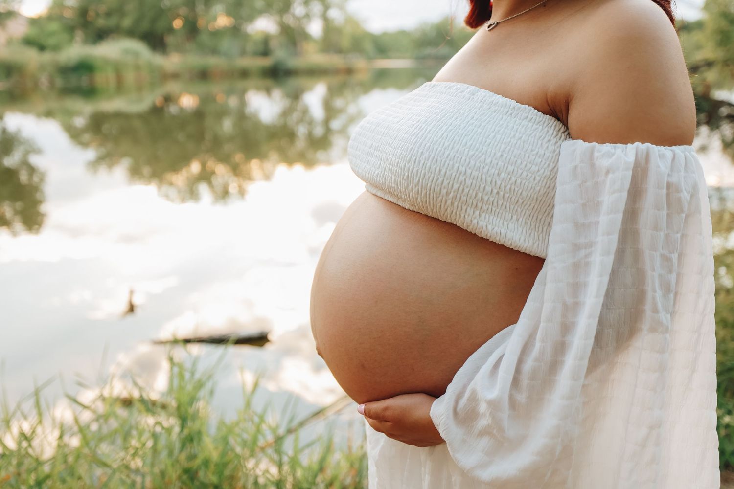 Close-up of pregnant woman in white off-shoulder top gently cradling bare baby bump near lake.