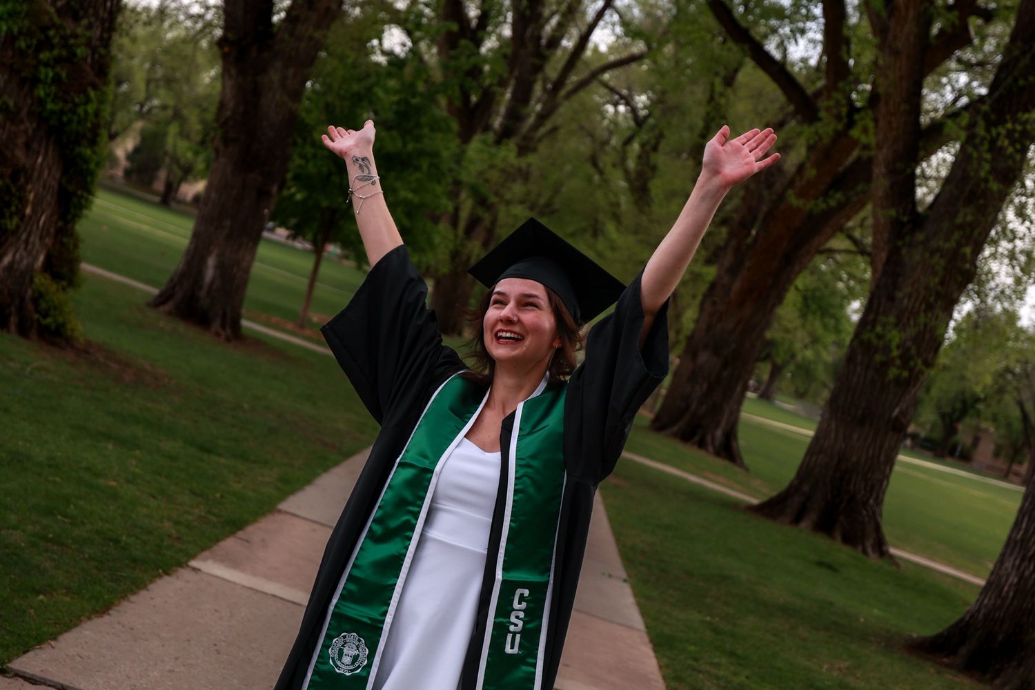 Joyful college graduate in black cap and gown with green stole celebrates outdoors with arms raised on tree-lined campus path.