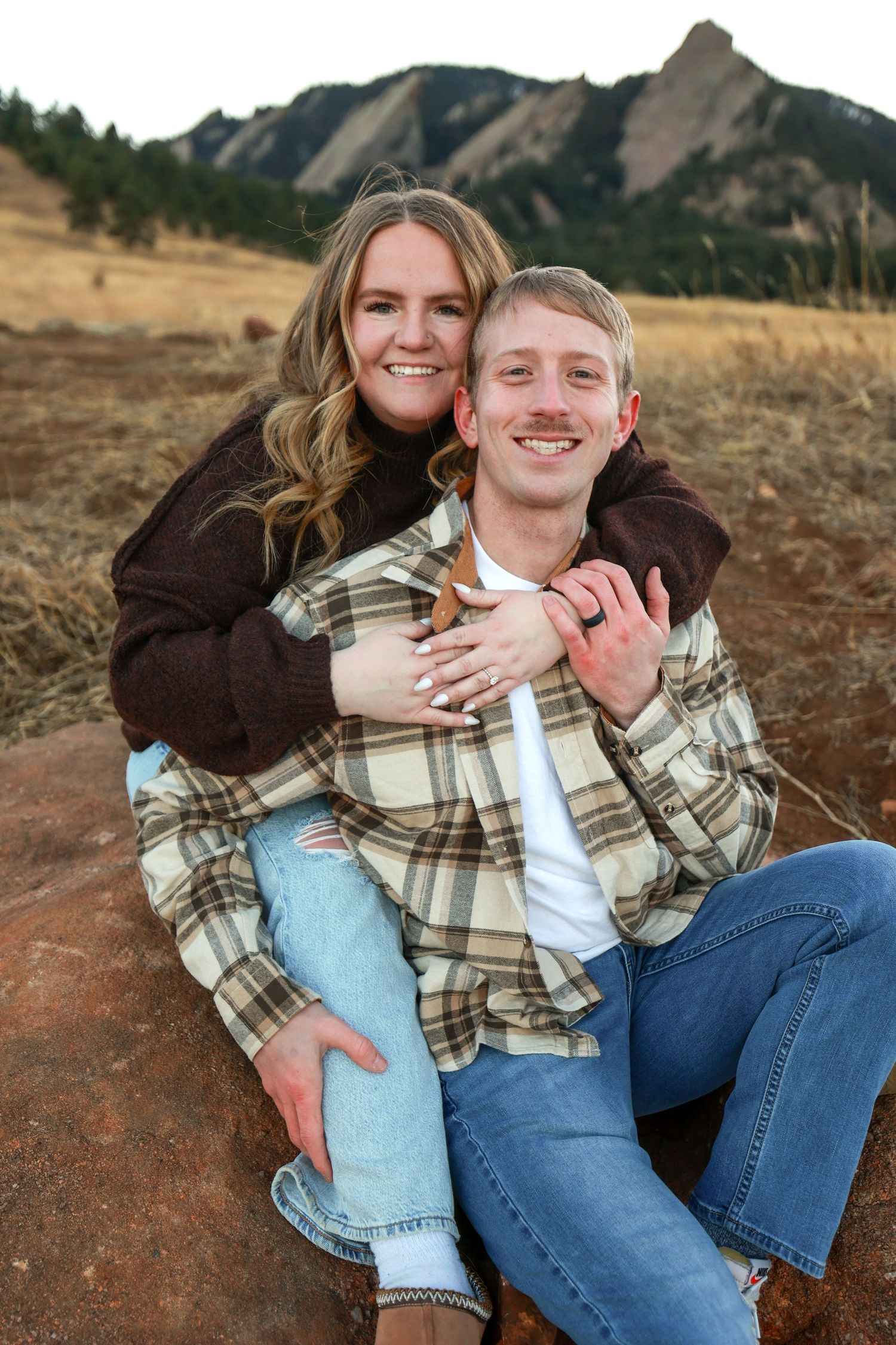 Smiling couple sitting on red rock with Colorado Flatiron mountains in the background.