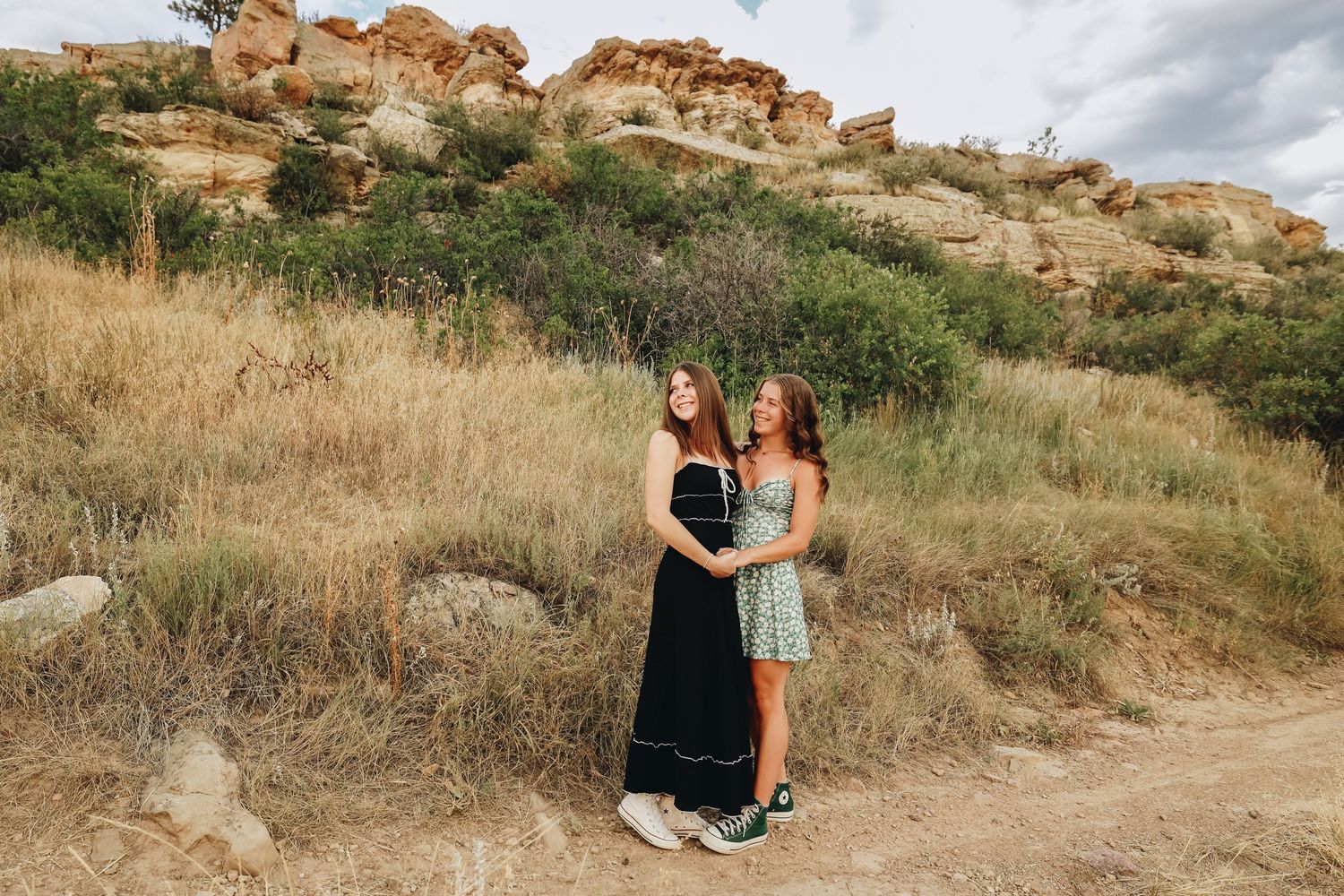 Two young women in summer dresses posing together on a dirt trail with rocky cliffs and dry grass.