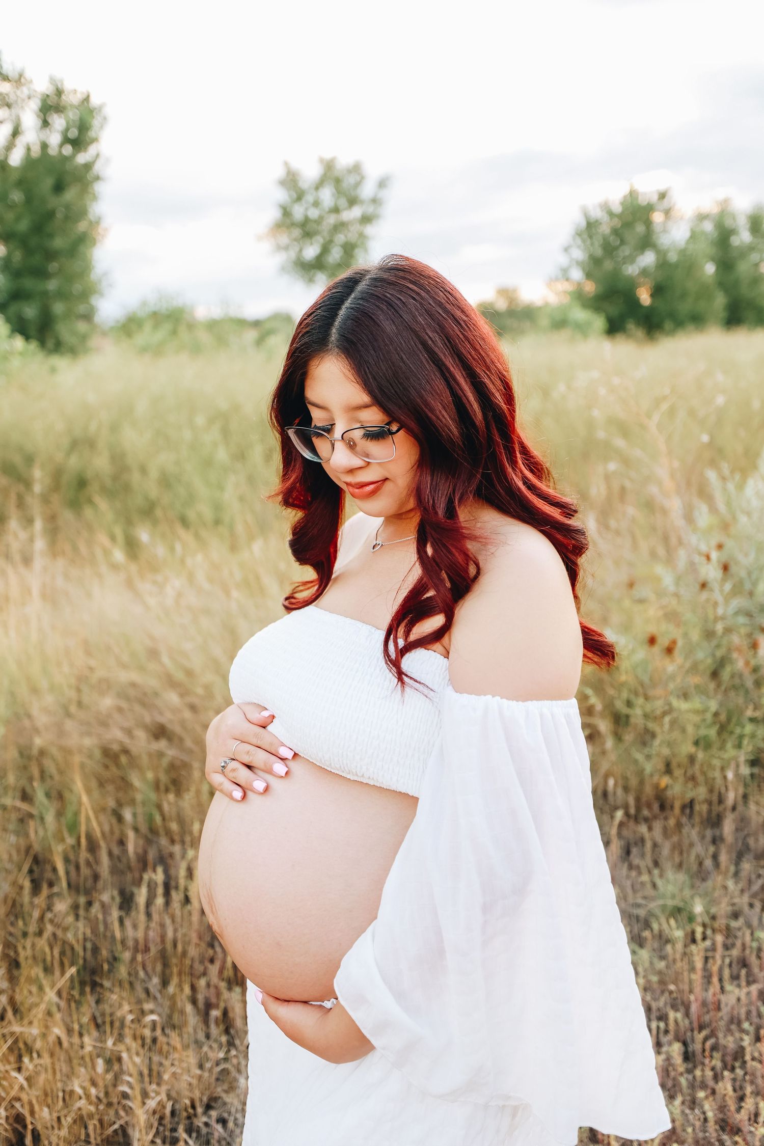 Pregnant woman in white off-shoulder dress cradling baby bump in golden field at sunset.