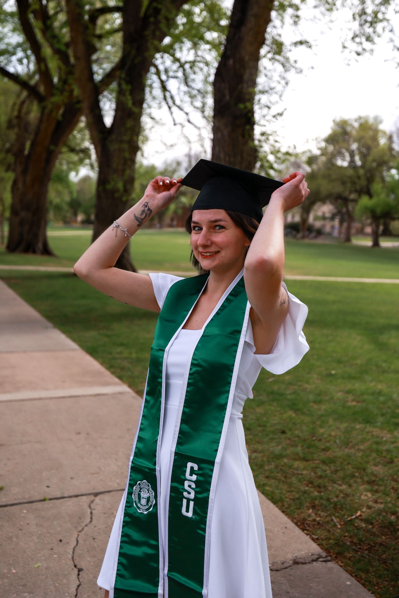 A smiling graduate in white gown and green CSU stole adjusts her black mortarboard cap outdoors on campus.