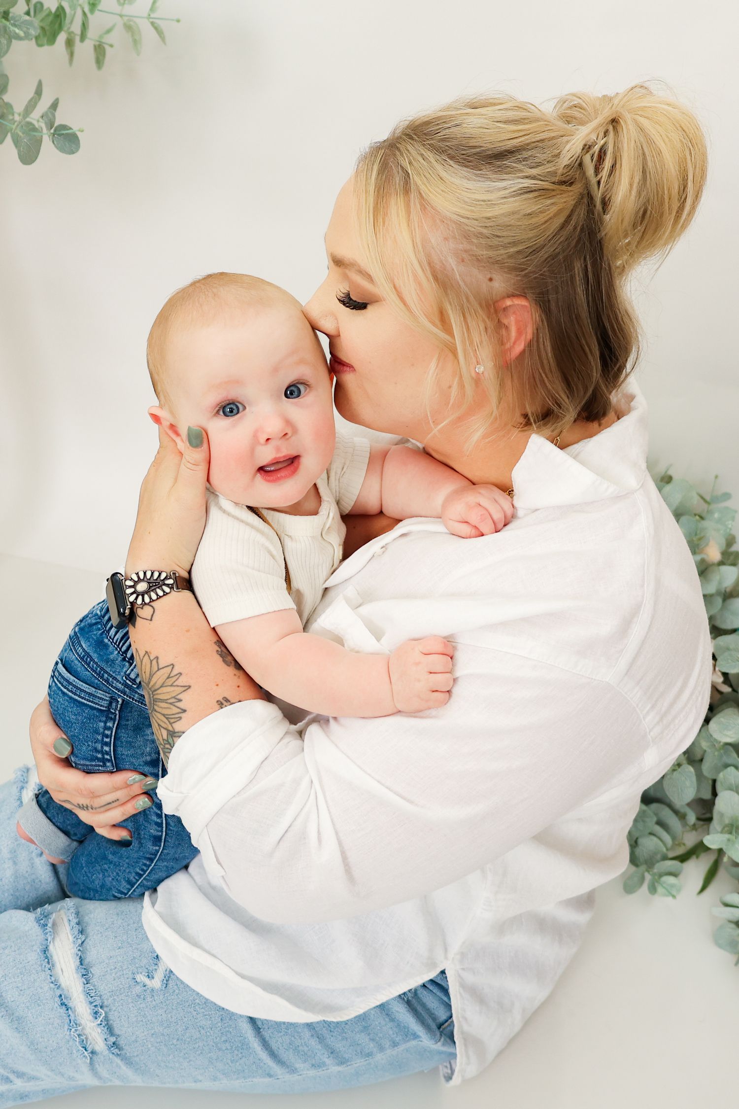 Blonde mother in white shirt and jeans kissing her blue-eyed baby during a bright studio photoshoot.