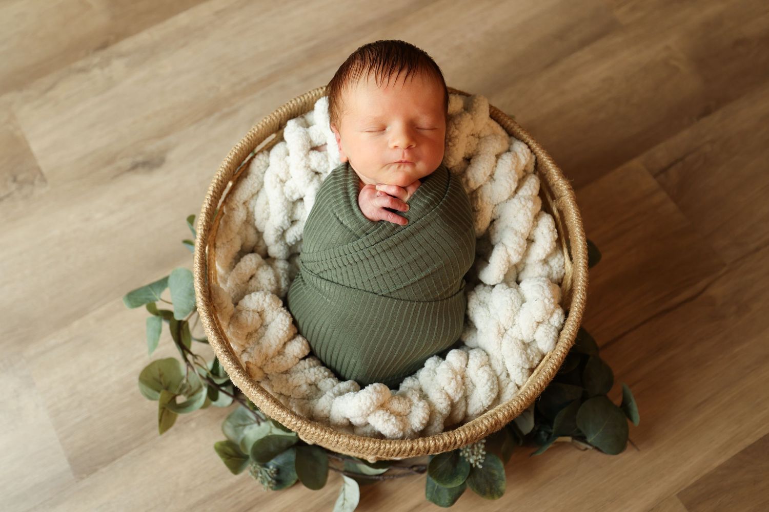Newborn baby boy swaddled in a green wrap sleeping in a wicker basket with eucalyptus.