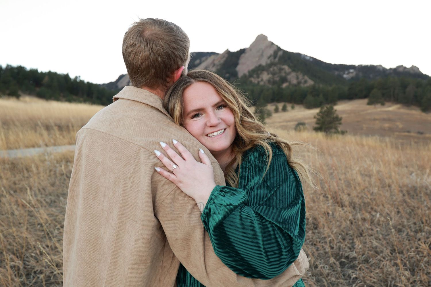Smiling woman hugging man from behind in golden grass field with mountain backdrop.
