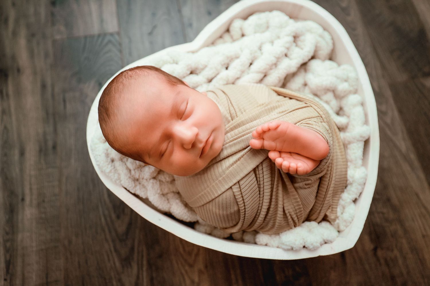 Sleeping newborn baby swaddled in beige wrap, nestled in a heart-shaped white bowl with knit blanket.