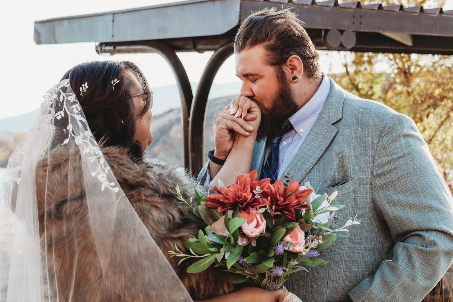 Groom kissing bride's hand while she holds a red and pink floral bouquet outdoors.