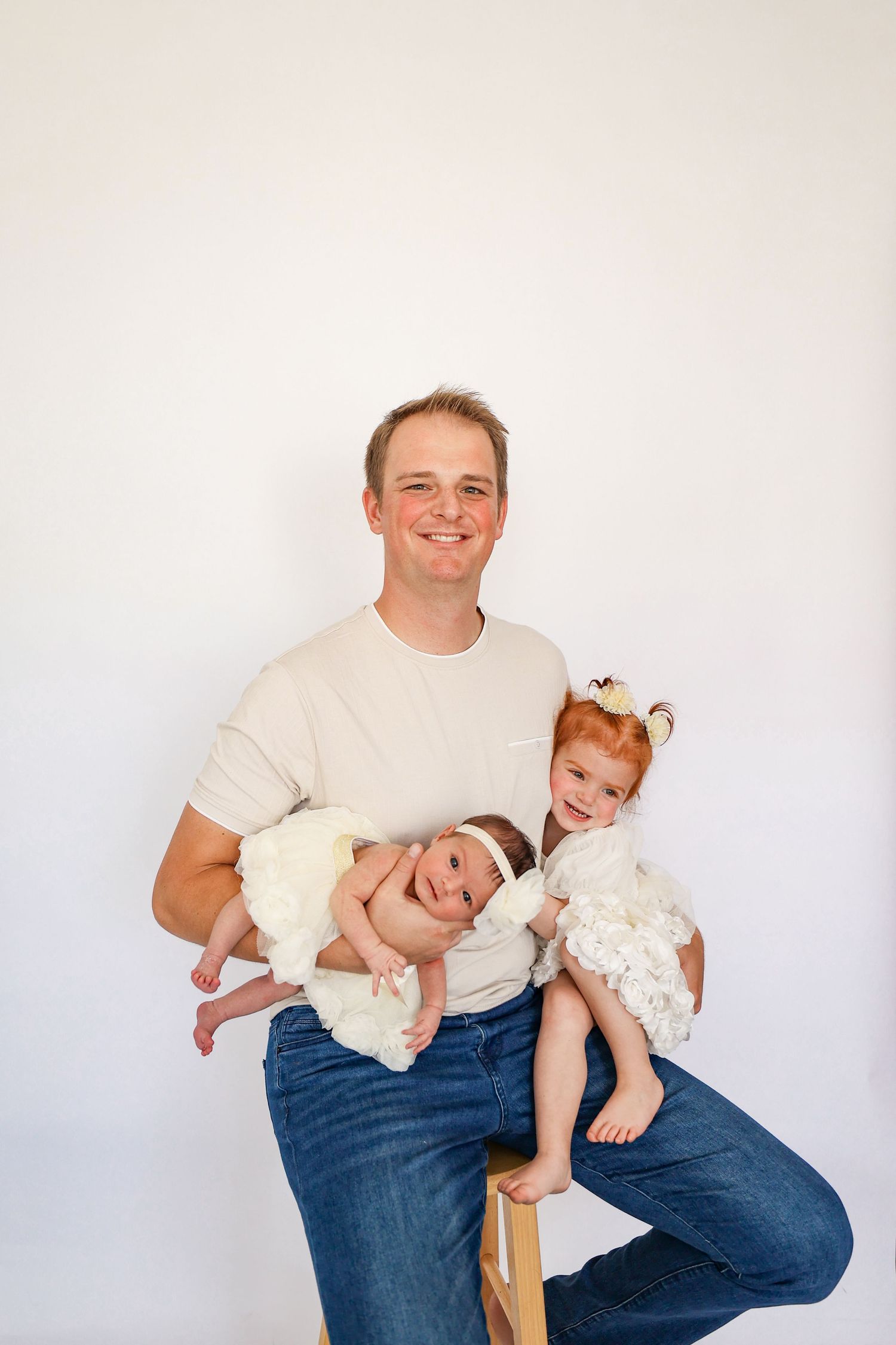 Proud father sitting and holding newborn baby and red-haired toddler daughter in white dress for portrait.