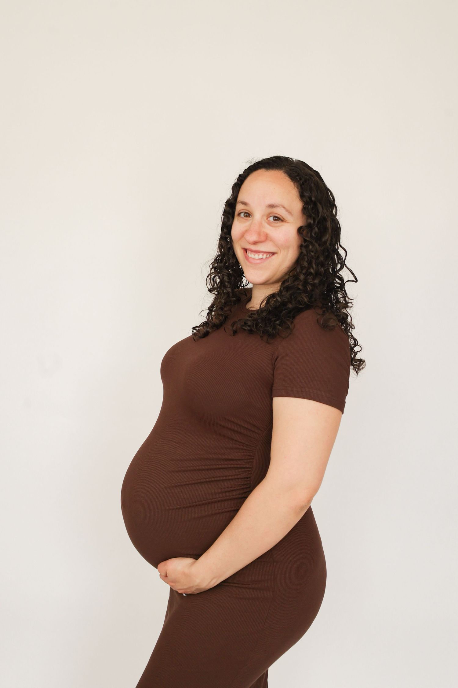 Smiling pregnant woman in brown dress cradling baby bump against white background.
