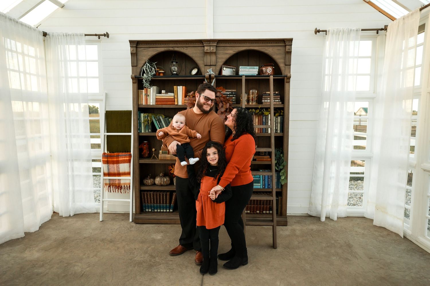 Family of four posing warmly in front of a large wooden bookshelf in a bright sunlit room.