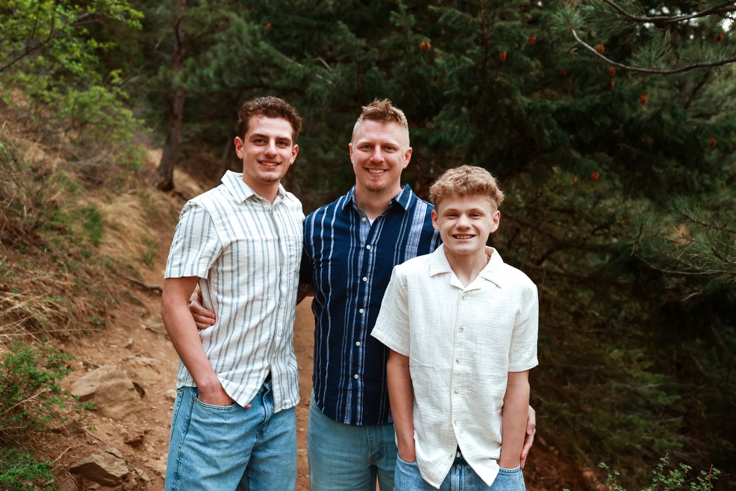 Three smiling young men pose together on a forest trail surrounded by lush green trees.