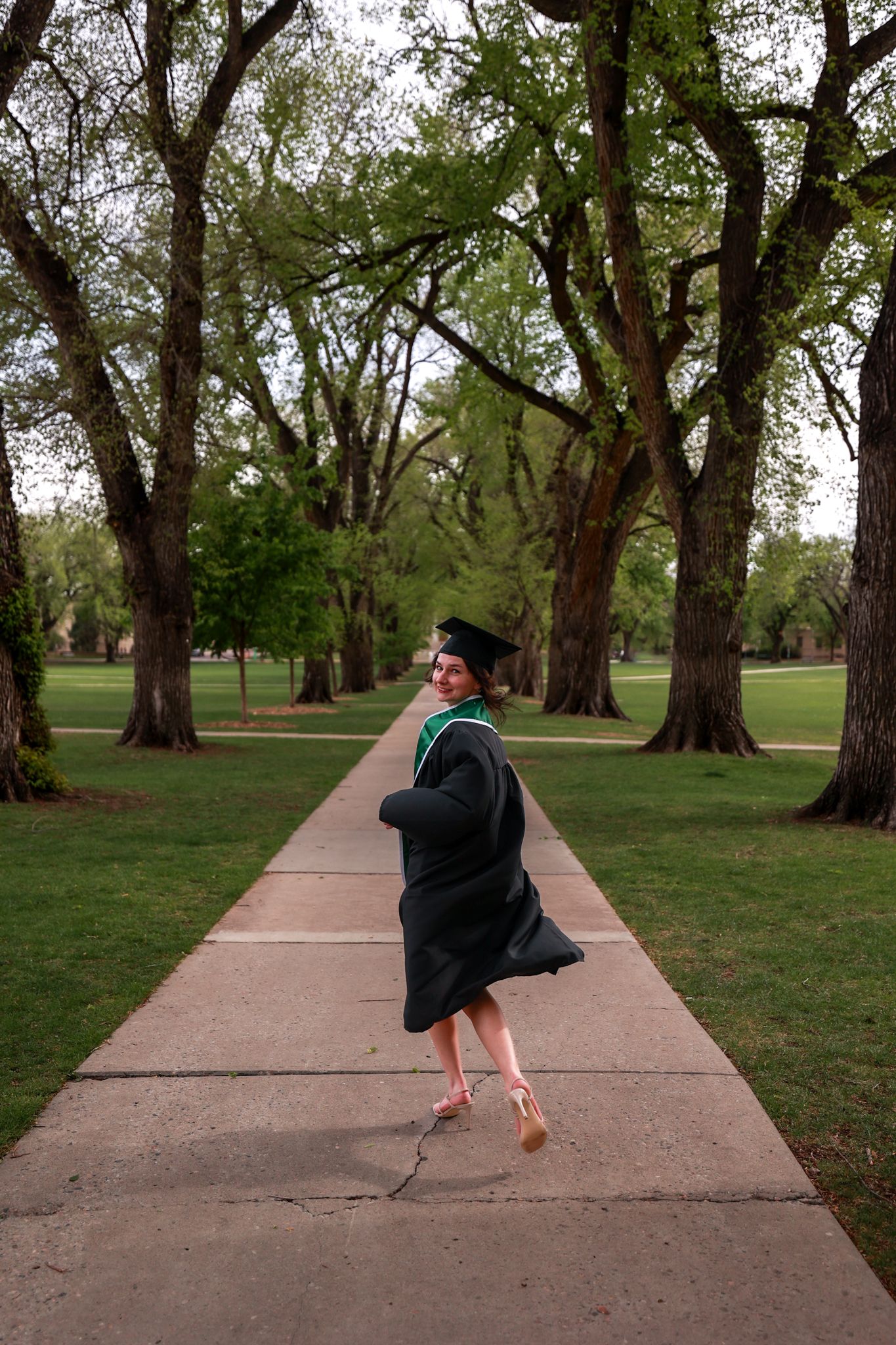 A joyful graduate in black cap and gown twirls on a tree-lined campus walkway, celebrating her graduation day.