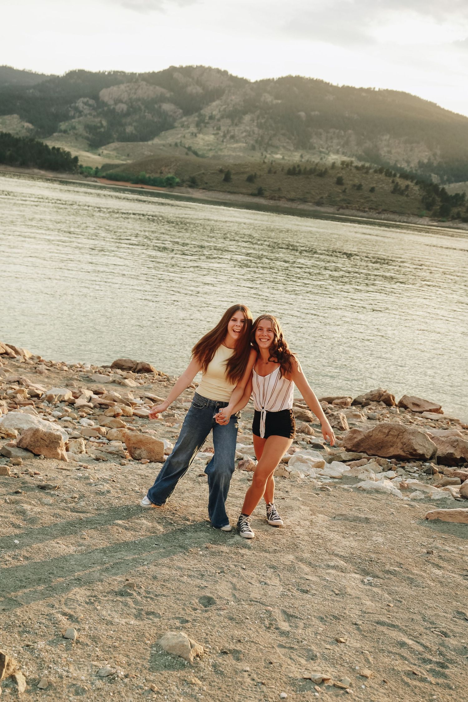 Two young women laughing and holding hands along rocky shoreline of mountain lake at golden hour.