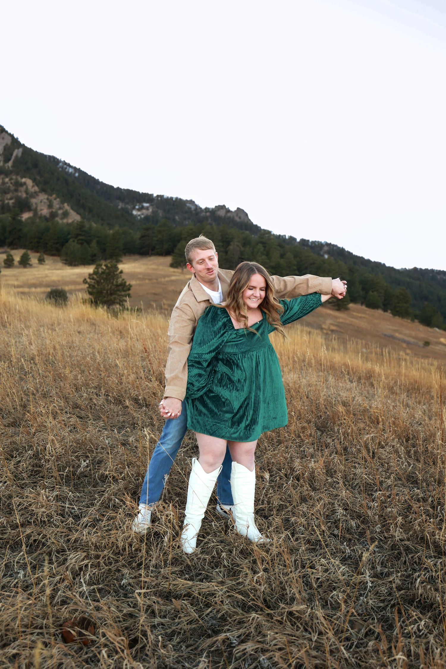 Joyful couple dancing in autumn field with rocky mountain backdrop, woman in emerald dress and white knee-high boots.