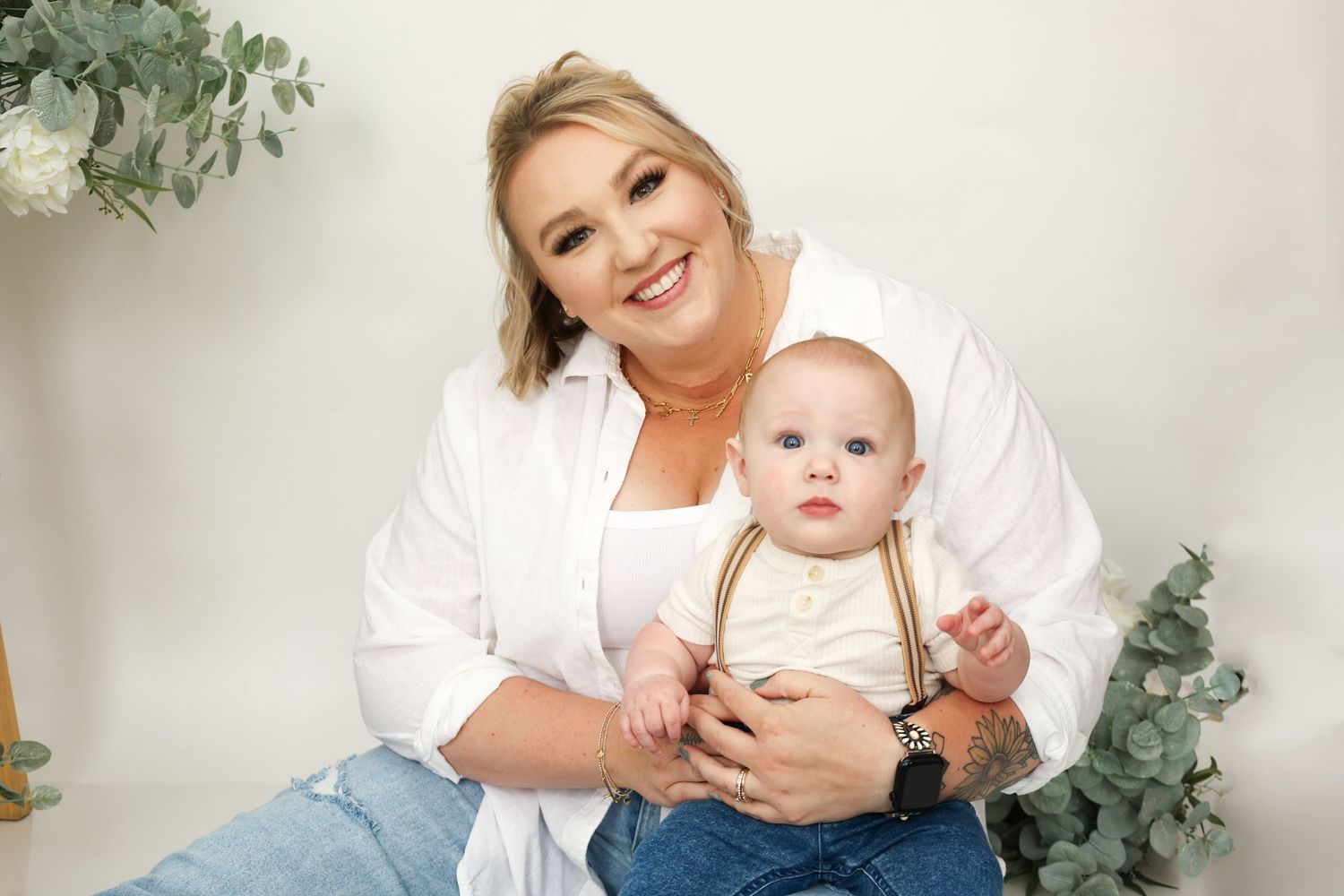 Cheerful blonde mother sitting with her baby boy in suspenders during a studio portrait session.