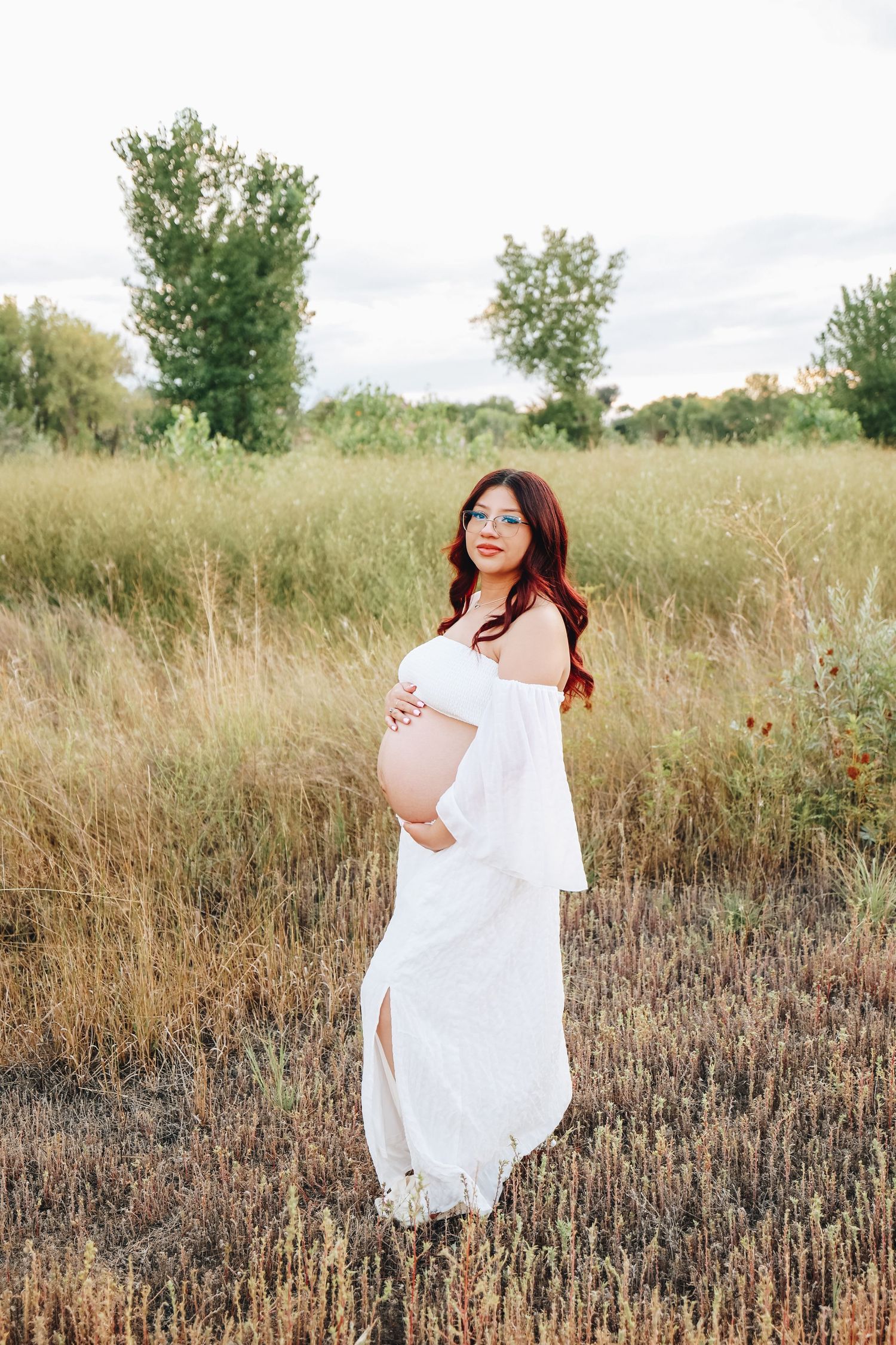 Pregnant woman in white off-shoulder dress holding belly standing in golden field at sunset.
