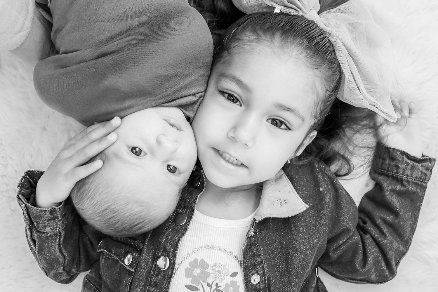 Black and white photo of two young siblings lying down, older girl smiling warmly while cradling baby sibling.