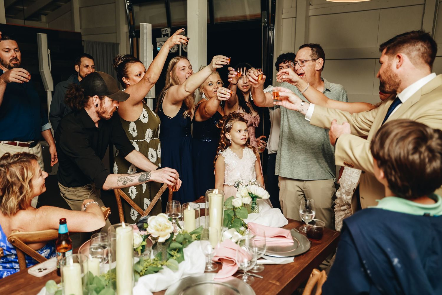 Wedding guests raising glasses in toast around flower-adorned reception table with young flower girl.
