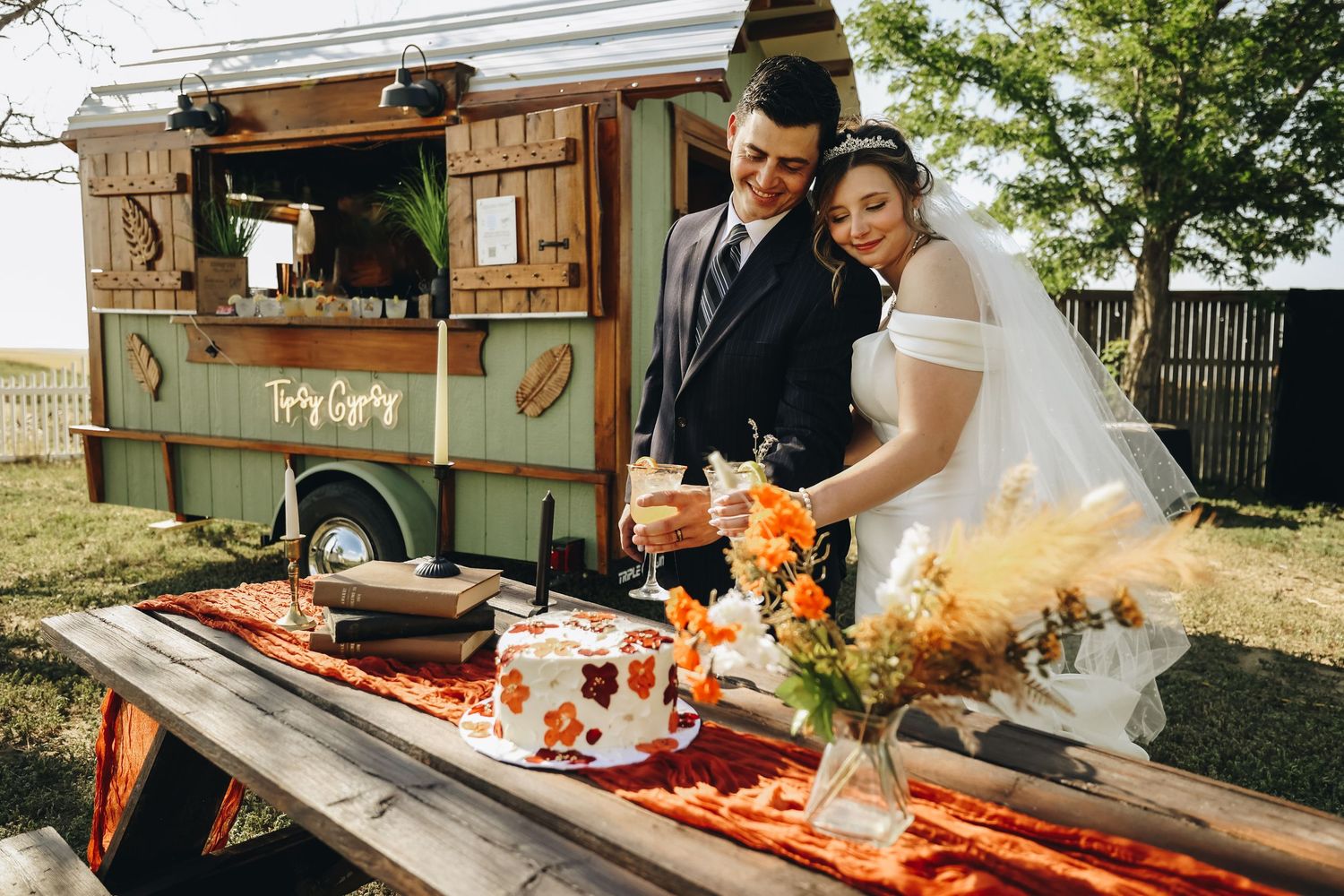 Bride and groom cutting their wedding cake at a rustic outdoor table beside a charming vintage food truck.