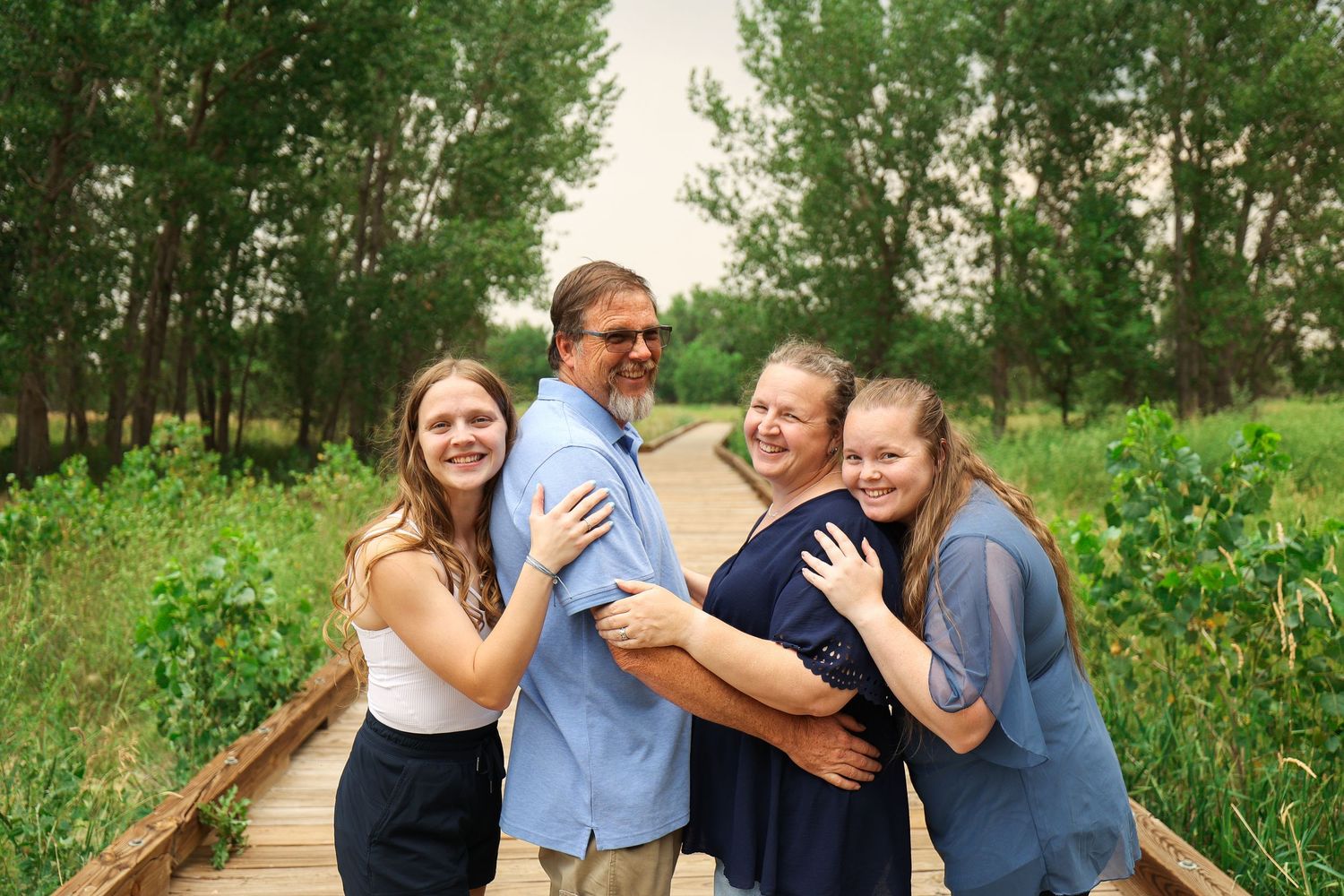 Happy family of four posing together on wooden boardwalk path in green nature setting.