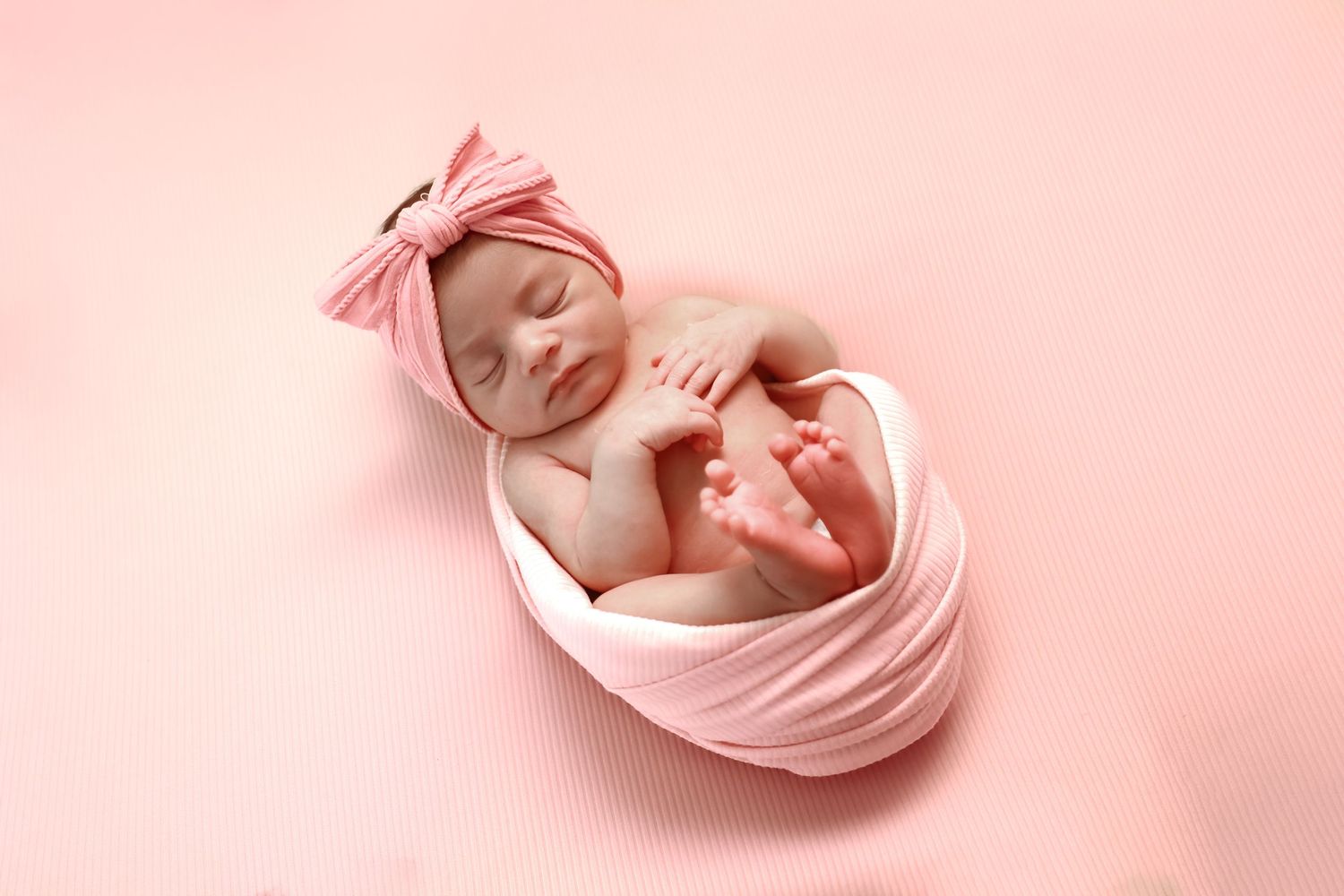 Newborn baby swaddled in soft pink wrap with bow headband sleeping peacefully on pink background.