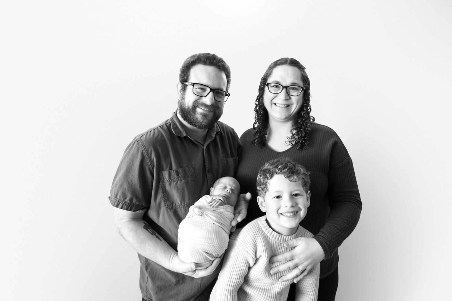 Black and white family portrait with parents holding newborn baby and young child smiling together.