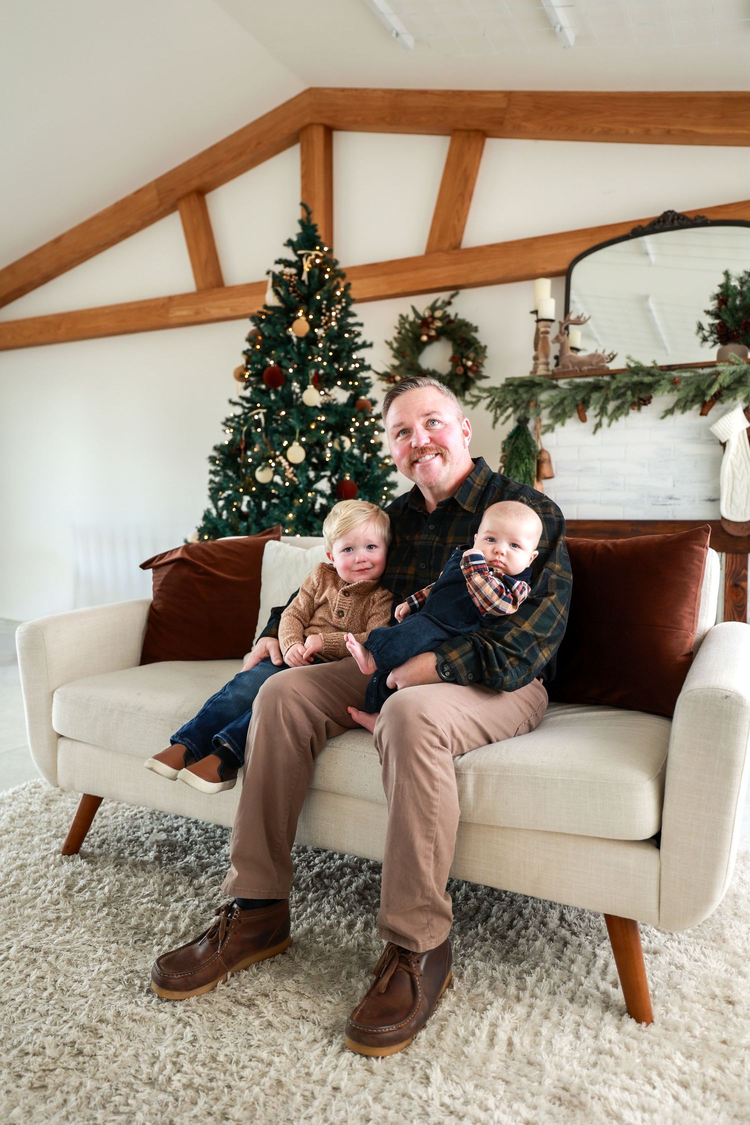 Smiling father sits on cream sofa holding toddler and baby in front of decorated Christmas tree at home.