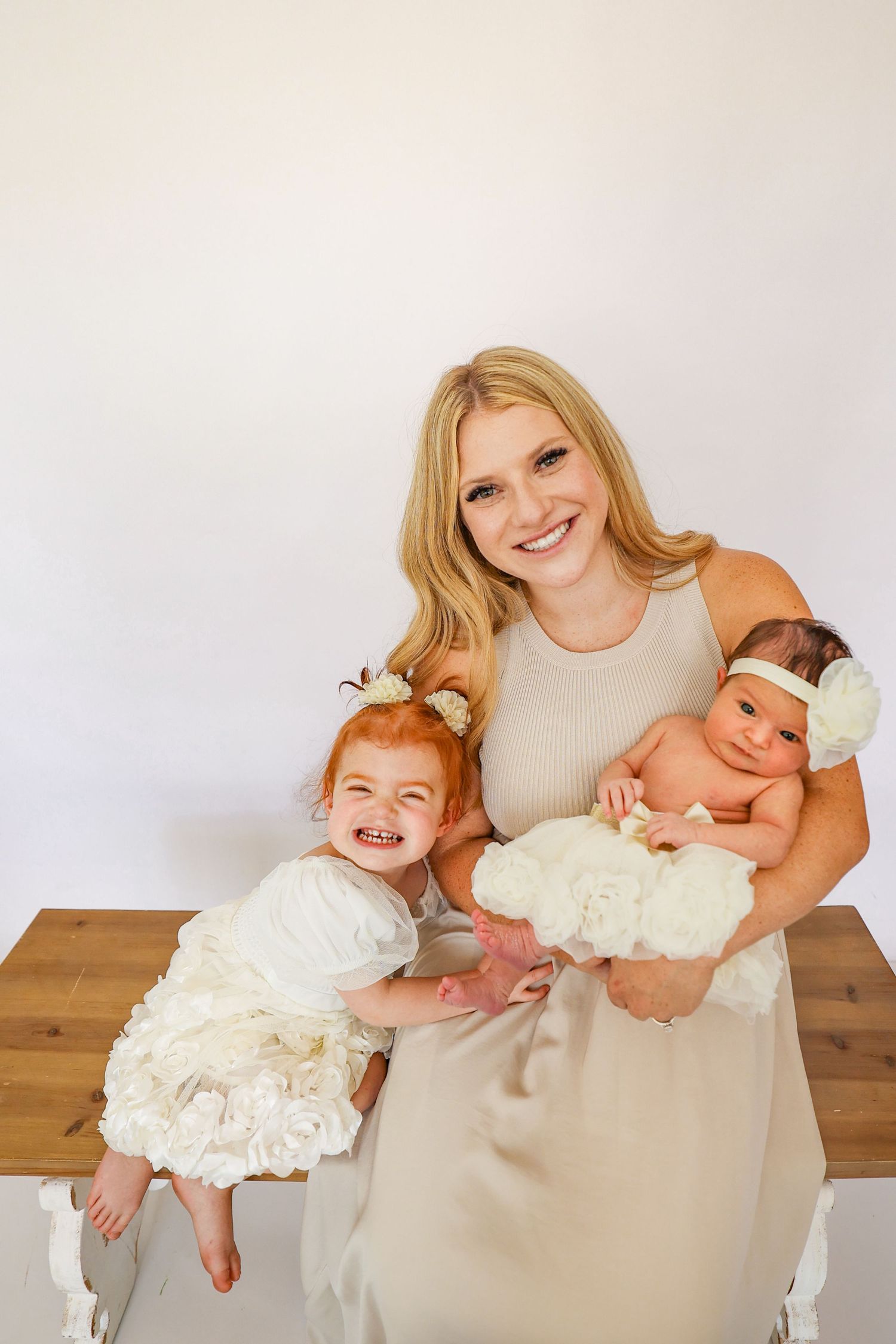 Smiling blonde mother holding newborn baby while toddler daughter in white dress poses for family portrait.