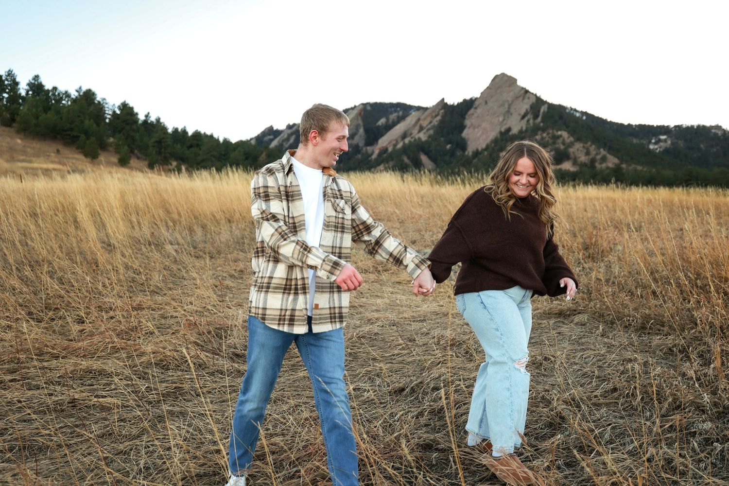 Happy couple holding hands and walking through dry golden field near Colorado Flatirons.