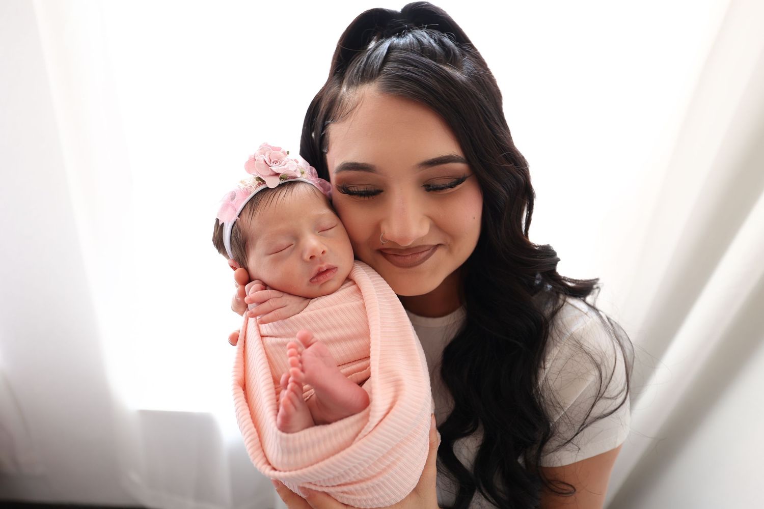 Smiling mother with long dark hair holding her sleeping pink-swaddled newborn baby girl.