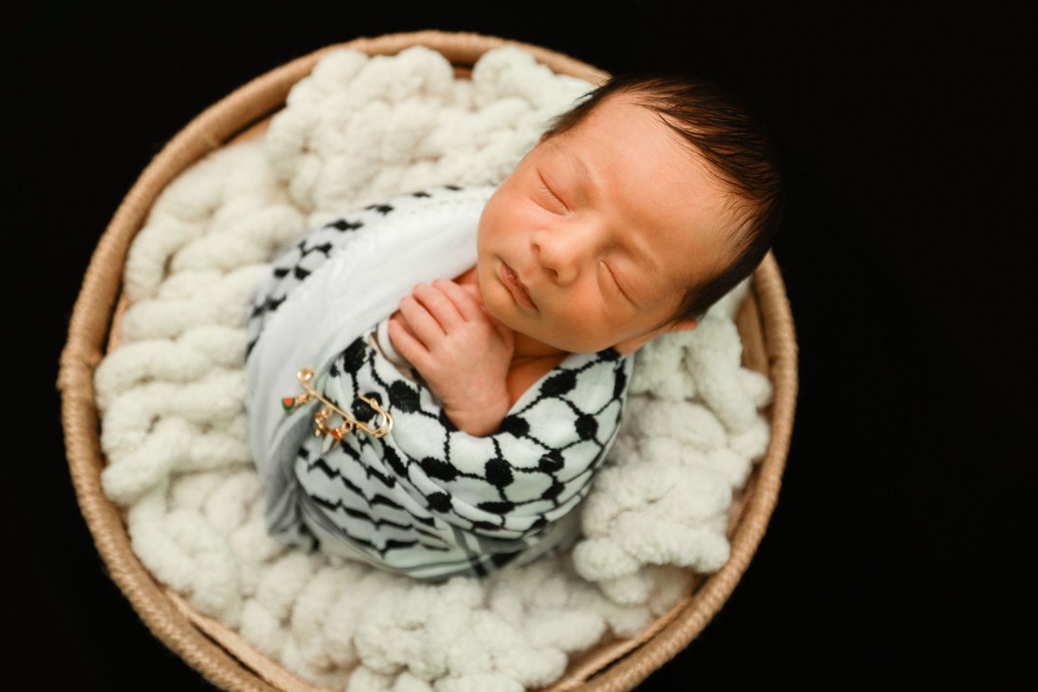 Sleeping newborn wrapped in keffiyeh pattern cloth resting in a wicker basket with white knit blanket.