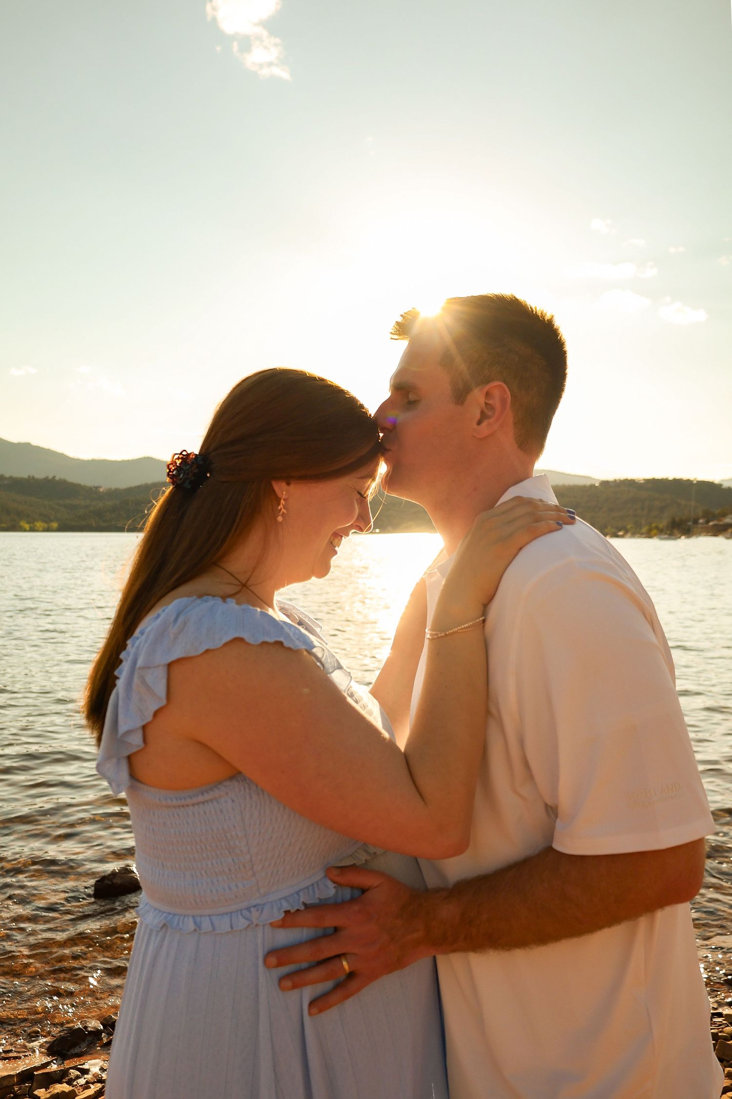 Pregnant couple sharing a kiss at sunset by a lake, woman in light blue maternity dress.