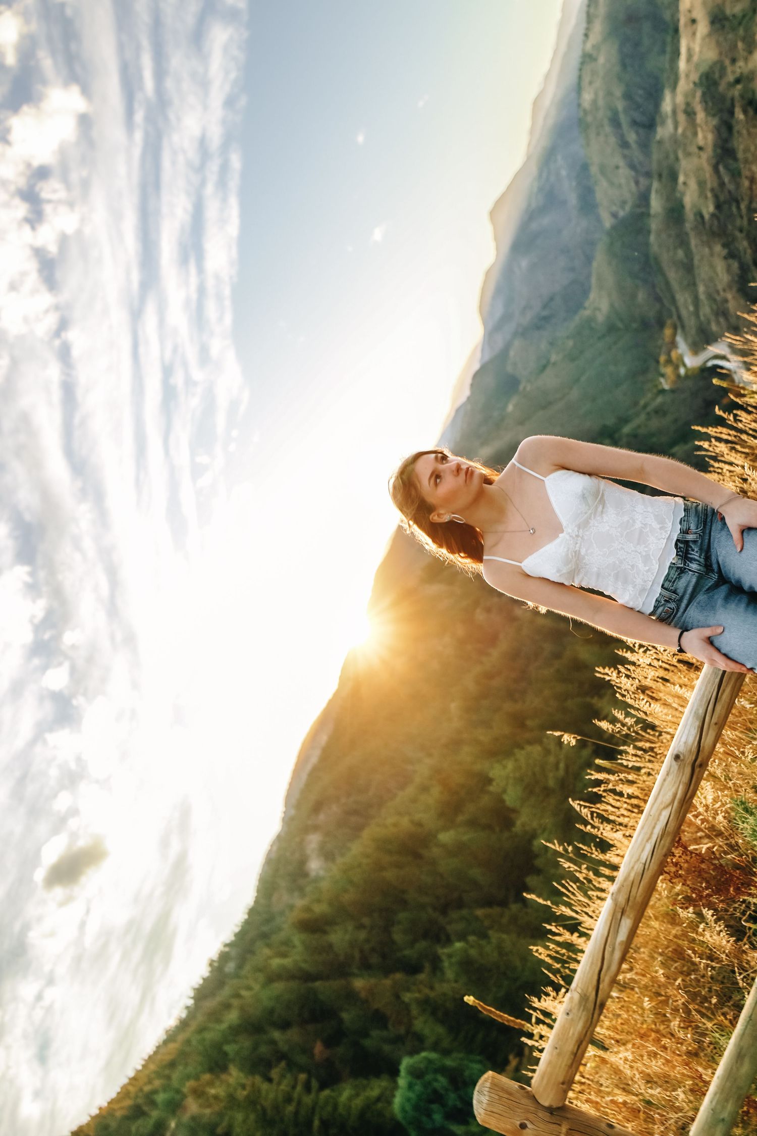 Young woman leaning on wooden fence railing with stunning mountain sunset landscape in background.