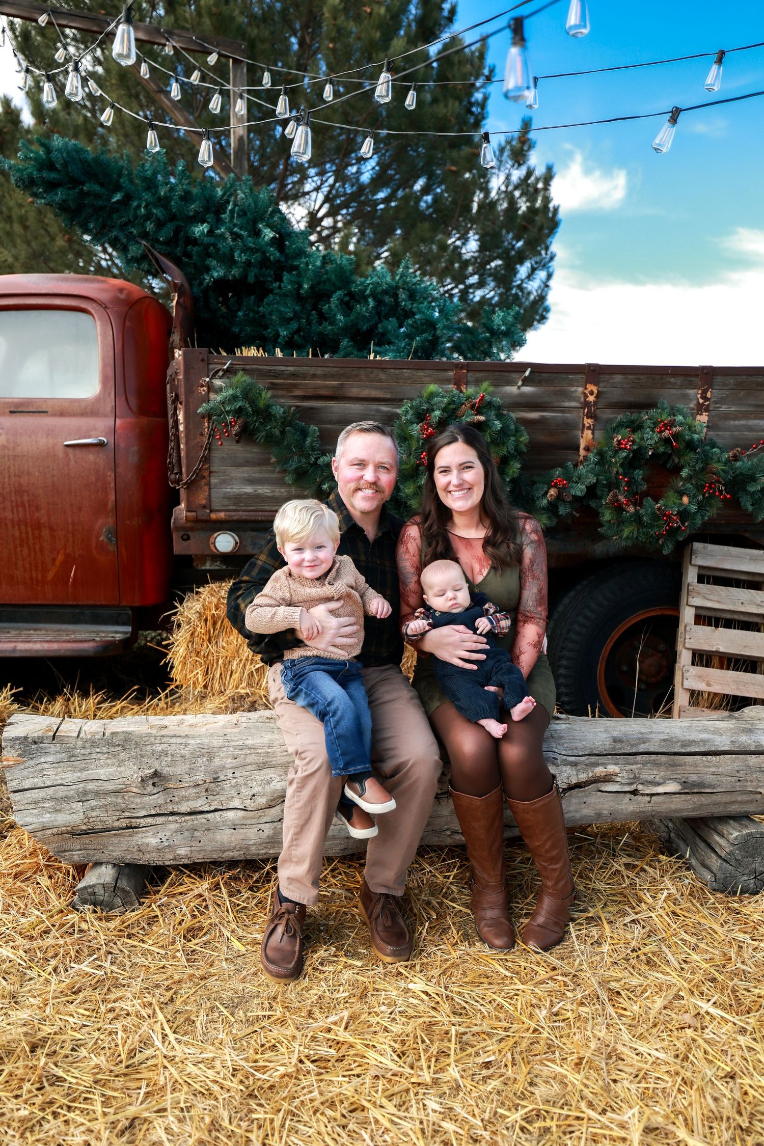 Family of four with two young children sit on log bales beside vintage red truck with Christmas wreaths outdoors.