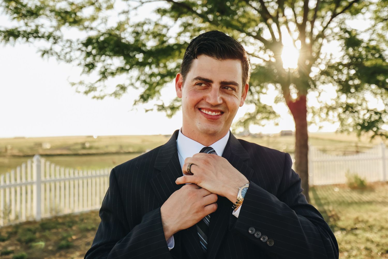 Handsome groom in navy pinstripe suit smiles while adjusting tie in warm golden sunlight outdoors.