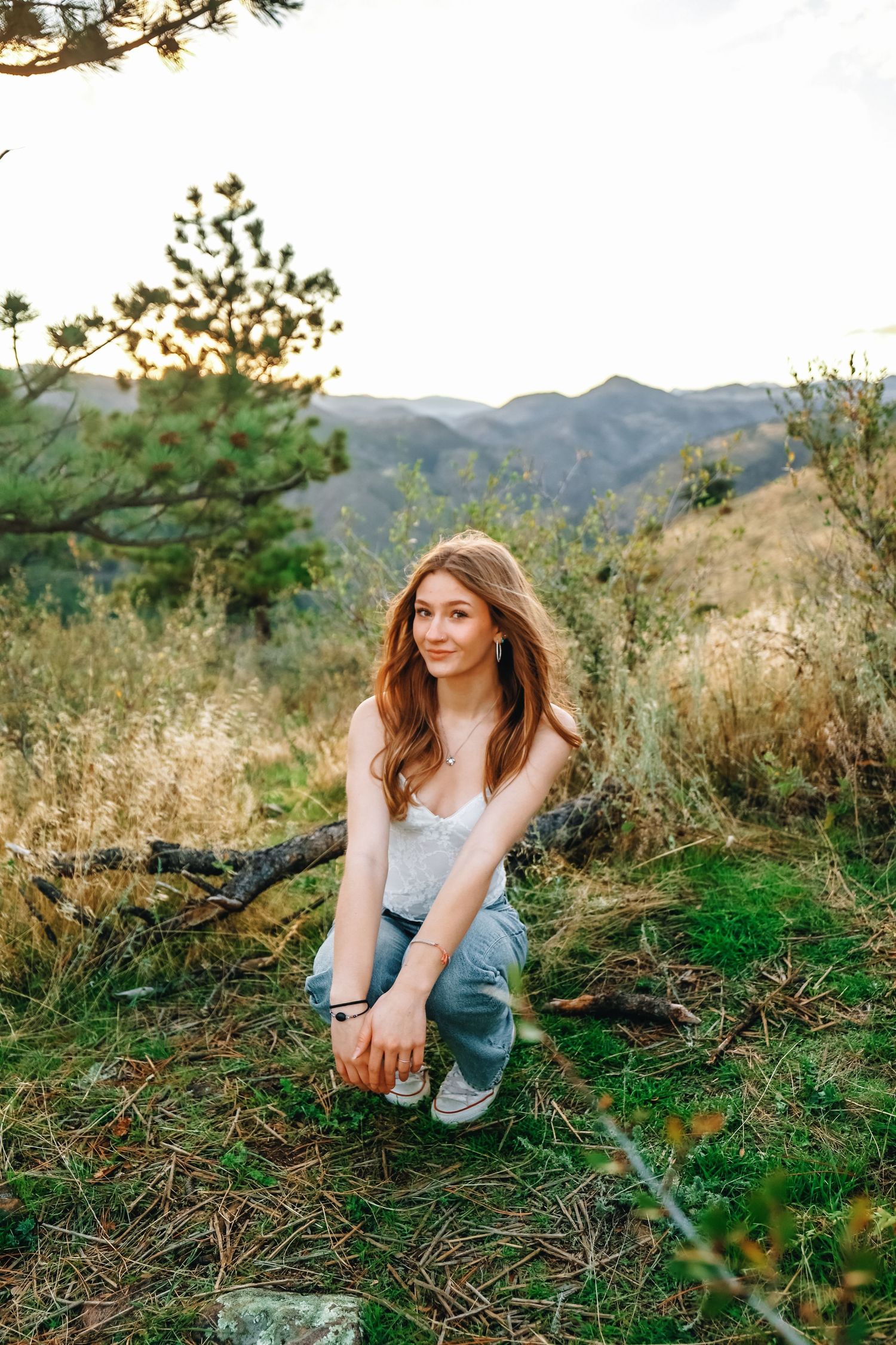 Young woman with long red hair in white top and jeans crouching in mountain meadow at golden hour.