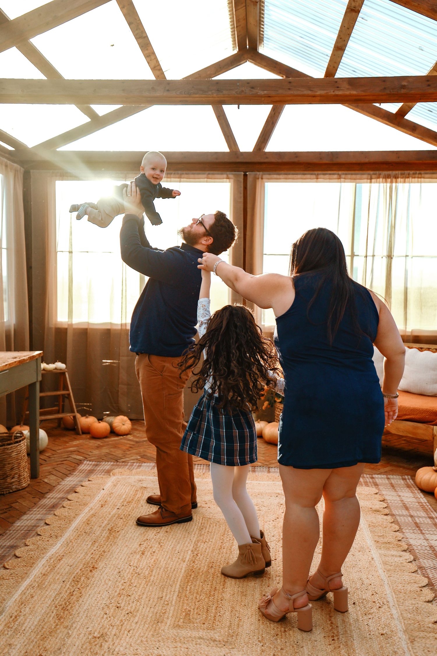 Family laughing together in a warm sunlit barn, father lifting baby in the air while mother and daughter watch joyfully.