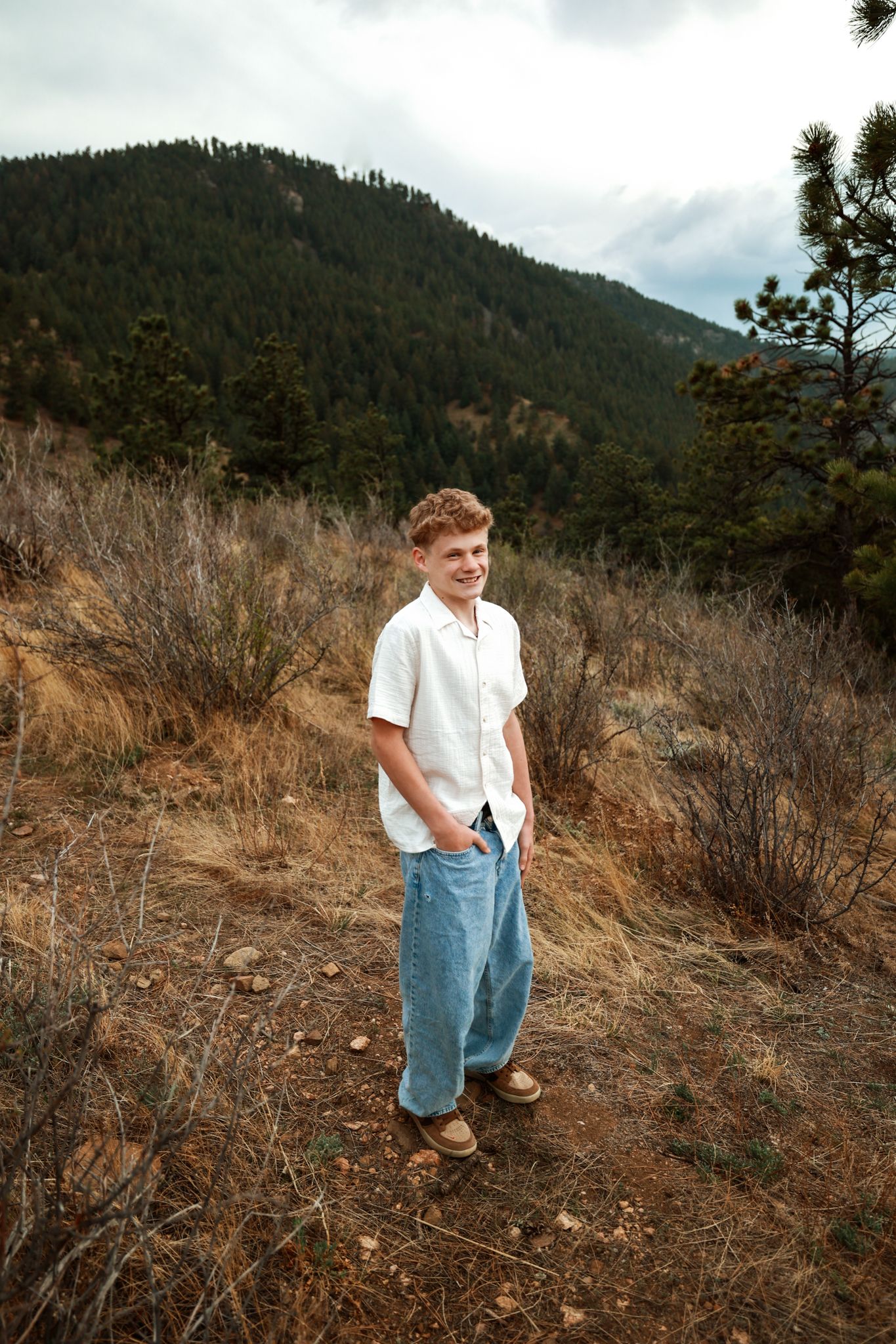 A young man in a white shirt and baggy jeans stands smiling on a mountain trail surrounded by pine trees.