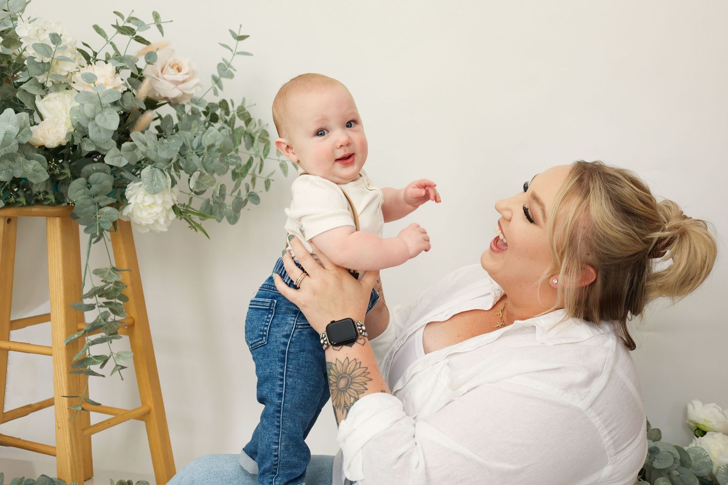 Blonde mother lifting and smiling at her happy baby boy near floral arrangements in a studio setting.