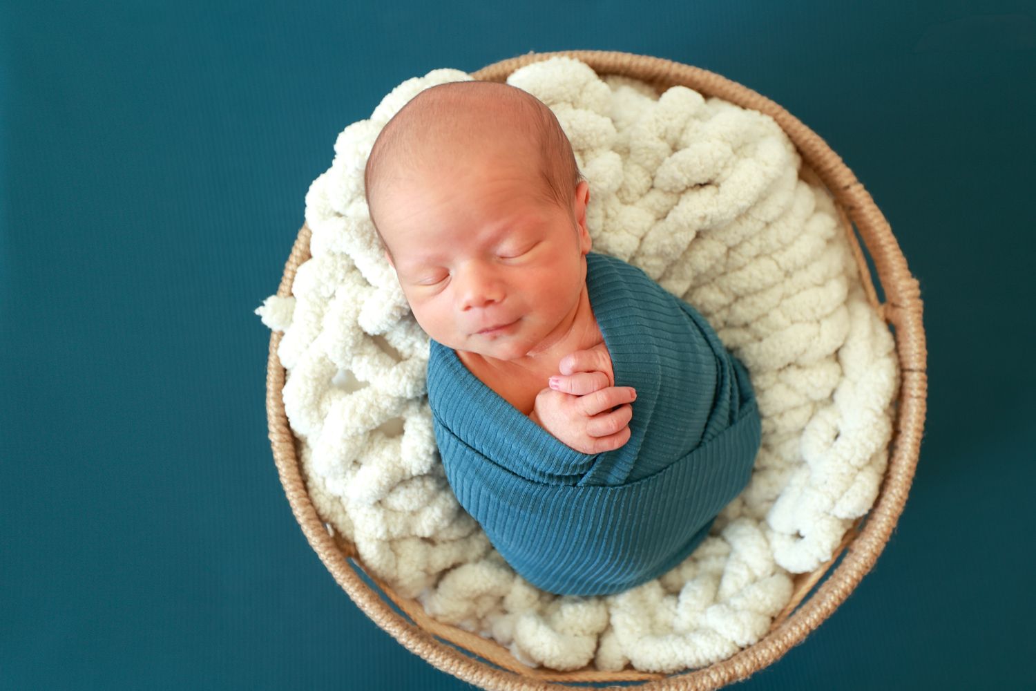 Peaceful newborn baby swaddled in teal wrap sleeping in a wicker basket with cream knit blanket.