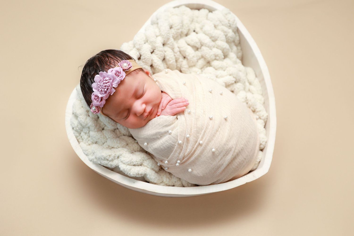 Newborn baby swaddled in cream wrap with pink floral headband sleeping in white heart-shaped bowl with knit blanket.