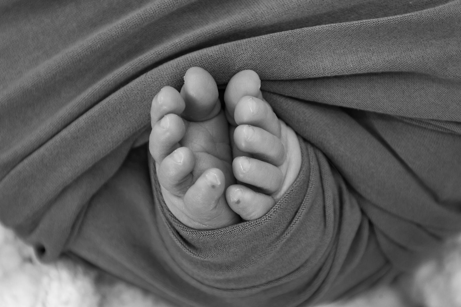 Black and white close-up of a swaddled newborn baby's tiny toes peeking through a soft gray wrap.