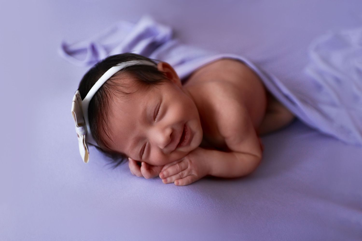 Smiling newborn baby girl in white headband sleeps peacefully on soft purple blanket in studio portrait.