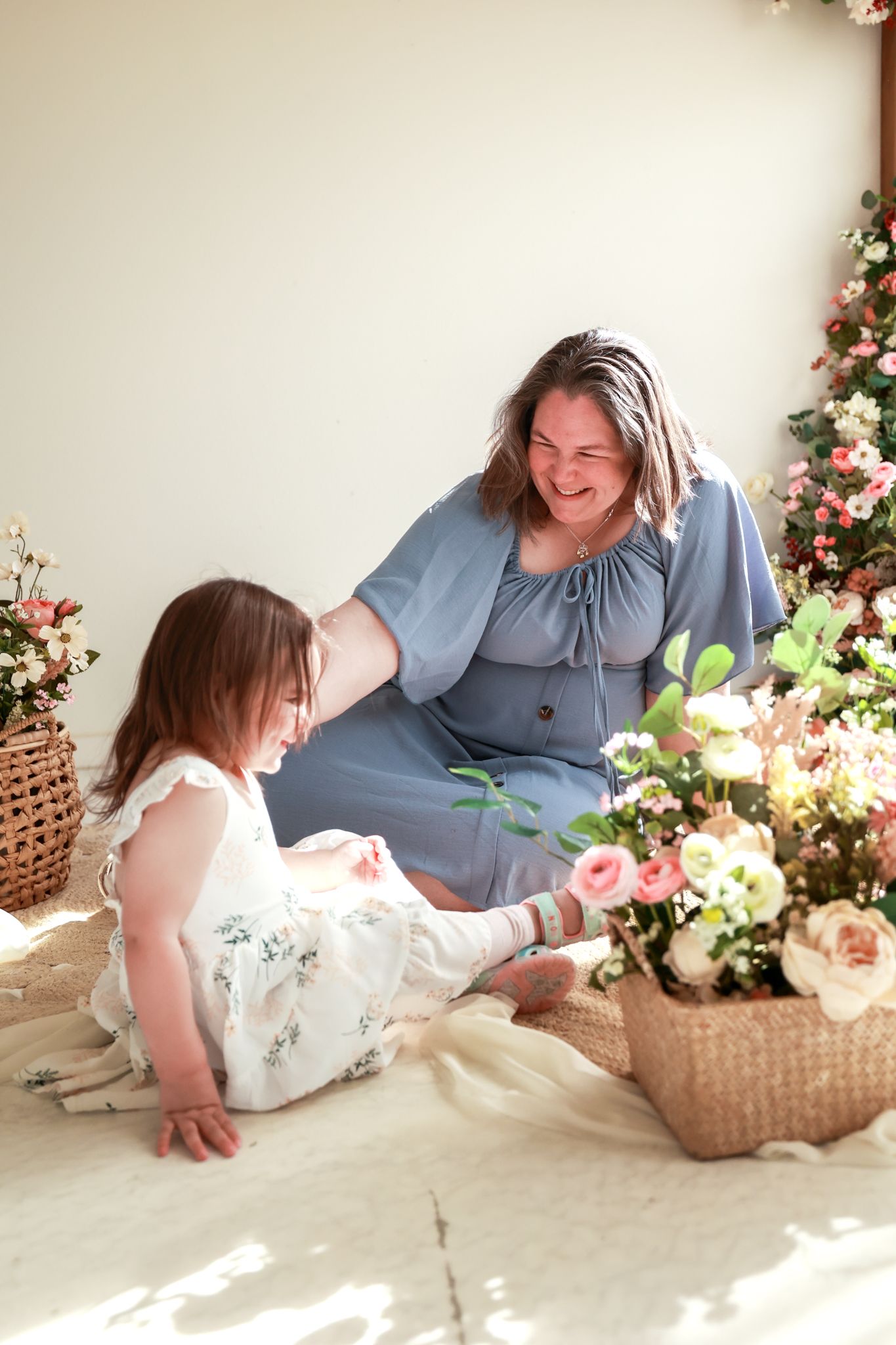 A woman in a blue dress laughs with a young girl in a floral dress surrounded by pink roses and flowers indoors.